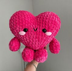 a pink heart shaped stuffed animal held up by someone's hand in front of a gray wall