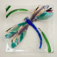 a glass dragonfly sitting on top of a white plate with green and blue feathers