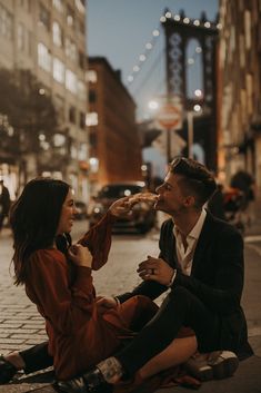 a man and woman eating pizza on the street at night with lights in the background