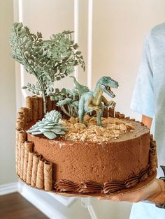 a person holding a cake decorated with dinosaur figurines and plants on top of it