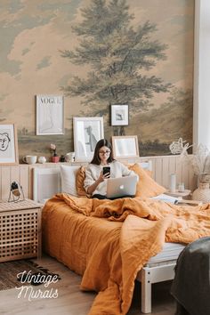 a woman is sitting on her bed with an orange comforter and holding a laptop