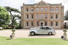 an old fashioned car parked in front of a large building with trees and bushes around it