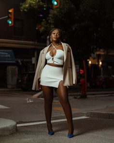 a woman is standing on the street at night wearing a white skirt and jacket with blue heels