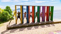 a group of people standing in front of a rainbow colored sculpture on the beach with water behind them