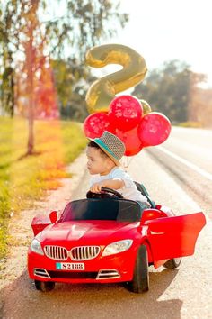 a little boy in a red car with balloons on the head and an s sign behind him