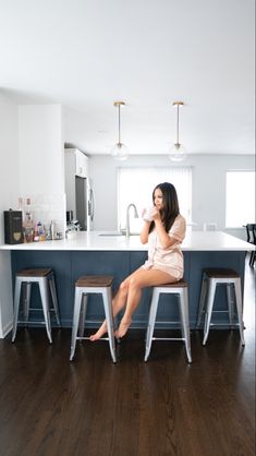 Long dark haired girl in satin champagne pajama shirt and shorts drinking coffee in a white cup sitting in front of a navy kitchen peninsula with white quartz countertop on a bar stool with dark wooden square top and 4 silver metal legs. Next to the girl are 3 of the same bar stool and her feet are on the legs of one of them. Above the kitchen peninsula is ceiling hanging brass pendant lighting with long vertical black wire and clear round glass. In the peninsula behind her is a sink Diy Kitchen Peninsula, Hale Navy Kitchen Cabinets, Hale Navy Kitchen, Kitchen Peninsula With Seating, Peninsula With Seating, Removing Wall, Small Kitchen Layout, Navy Kitchen Cabinets, Modern Shaker Kitchen