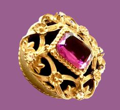 a gold ring with a pink stone in the center and ornate designs around it on a purple background