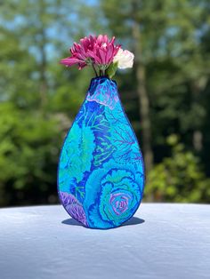 a blue vase with pink and white flowers in it on a table outside near some trees