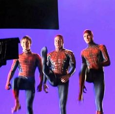 three people dressed as spider - man in front of a purple background, one holding a camera