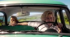 two women sitting in the driver's seat of a green car, one driving