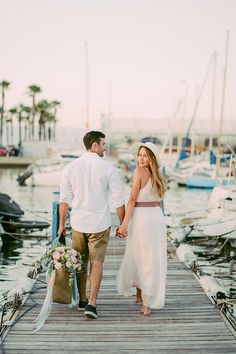 a man and woman holding hands walking on a dock with boats in the water behind them