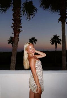 a woman standing next to two palm trees at night with her hand on her head