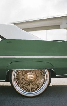 an old green car with chrome rims parked on the side of the road in front of a bridge