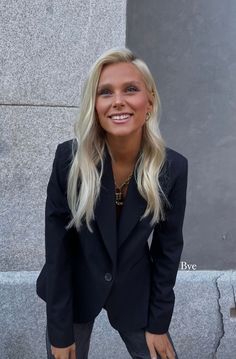 a woman with long blonde hair wearing a black blazer and jeans, standing in front of a wall