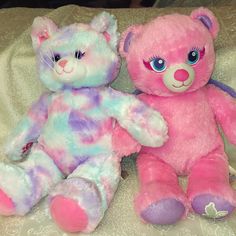 two pink and blue teddy bears sitting next to each other on a white bed sheet