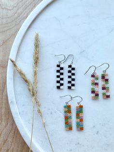 three pairs of beaded earrings sitting on top of a white plate next to a dried plant