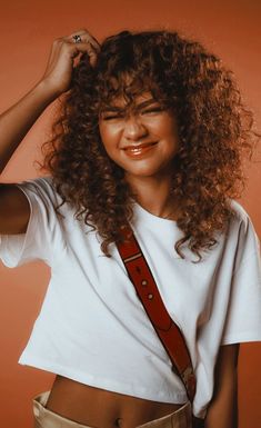 a woman with curly hair wearing a white t - shirt and tan pants is smiling