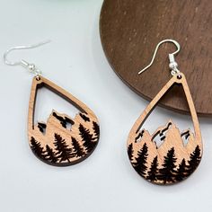 a pair of wooden earrings with mountains and trees on them sitting next to a piece of wood