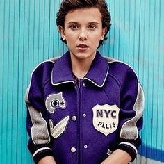 a young boy in a purple jacket is leaning against a blue wall with his hands on his hips