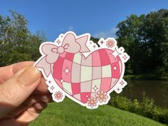 a person holding up a pink heart shaped sticker in front of a river and trees