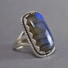 SKU: SS-013Material: 925 stamped sterling silverGemstone(s): Labradorite (N)Gemstone Dimensions: 12 X 22 X 3 mmRing Size: US 7Weight: 7.9 gramsThis gorgeous Labradorite cabochon ring features a large rectangular Labradorite cabochon, set in a wide jagged edge bezel and accented with a braid border. The Labradorite glows with an amazing blue-green fire, and it has a layer of mica that makes it glow AND shimmer at the same time. A truly magnificent stone! Silver Labradorite Hand Forged Rings, Hand Forged Silver Labradorite Rings, Luxury Silver Labradorite Rings, Jagged Edge, Luxury Cabochon Labradorite Rings, Green Fire, Silver Labradorite Cabochon Gemstones, Dendritic Agate, Cabochon Ring