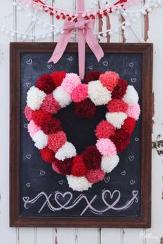 a valentine's day wreath hanging on a chalkboard with pom - poms