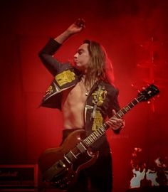 a man with long hair and no shirt on holding a guitar in his right hand