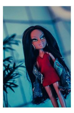 a doll with long black hair wearing a red dress and denim jacket is standing next to a plant