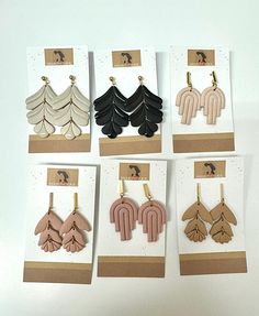 six pairs of earrings are hanging from hooks on a white surface, each with different shapes and colors