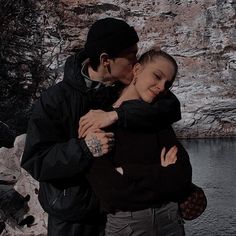 a man and woman embracing each other in front of a body of water with rocks behind them