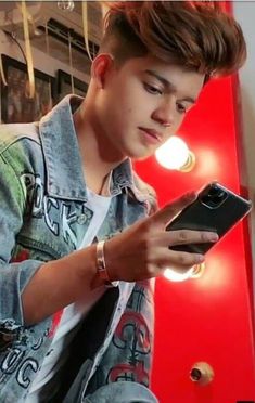 a young man sitting in front of a red wall looking at his cell phone while wearing a denim jacket