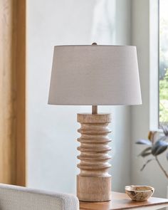 a table lamp sitting on top of a wooden table next to a white chair and window