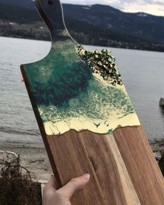 a hand holding a wooden board with an ocean scene painted on it and mountains in the background