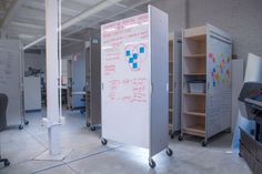 a whiteboard with writing on it is in the middle of an open room, surrounded by shelves and chairs