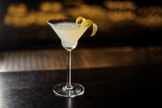 a white cocktail with lemon garnish on the rim sits on a black table