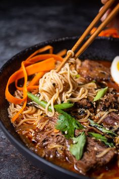 someone holding chopsticks over a bowl of noodles with beef and egg in it