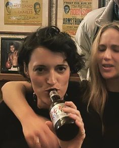 two women are sitting down and drinking from beer bottles in front of them, while another woman looks on