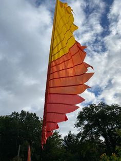 a large yellow and red kite flying in the air with trees behind it on a cloudy day