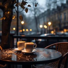 two cups of coffee sitting on top of a wooden table next to a tree in the rain