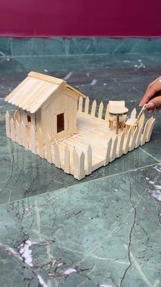 a wooden model of a house with a fence on it's sides and a hand reaching for the door