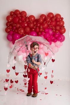 a young boy holding an umbrella in front of balloons and confetti hearts on the wall