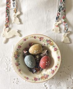 three easter eggs in a bowl on a doily with bunny decorations around them and two bunnies hanging from the ceiling