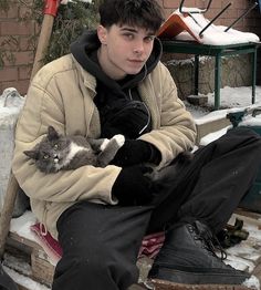 a young man sitting in the snow holding a gray and white cat on his lap