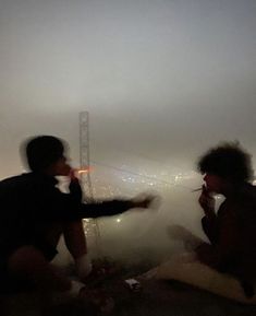 two people sitting on the ground in front of a foggy cityscape at night