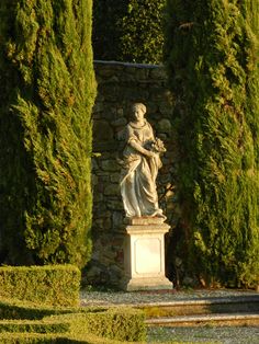 a statue in the middle of a garden surrounded by trees and hedges, with a stone wall behind it