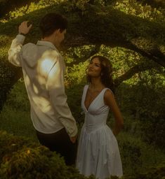 a man standing next to a woman in a white dress under a tree with the sun shining on her