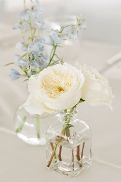 two clear vases filled with white and blue flowers on top of a tablecloth