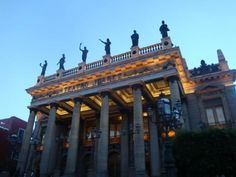 an old building with columns and statues on the roof at dusk, lit up by street lights