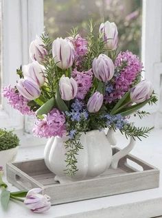 a white pitcher filled with purple flowers sitting on top of a table next to a window