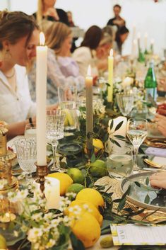 people sitting at a long table with plates and glasses on it, surrounded by candles